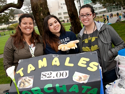 Three individuals sitting at a table selling tamales for two dollars