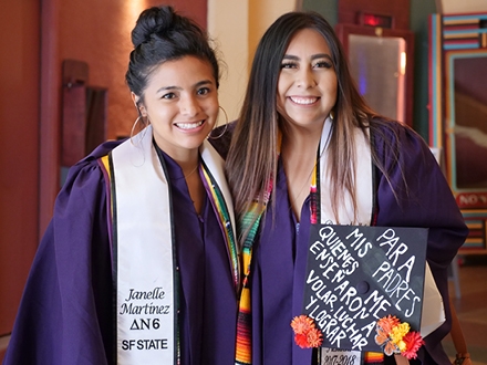 Two students in their graduation gowns with one student holding their graduation cap that is decorated. 